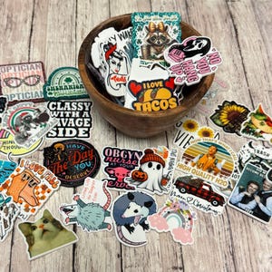 May include: A collection of assorted vinyl stickers featuring various graphic designs, text, and illustrations, including a raccoon, a ghost, a cat, and a truck, with many stickers contained within a round wooden bowl and others scattered around.
