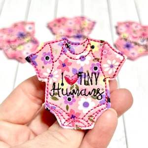 May include: Pink floral baby onesie applique with the text "I ❤️ TINY HUMANS" in black script. The applique has a red stitched outline and is decorated with purple, yellow, and white flowers. Several similar appliques are in the background.