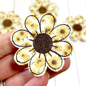 May include: Close-up of a sunflower-shaped applique with a brown center and yellow petals. The petals are decorated with a sunflower pattern. The applique has a black stitched outline. Other similar appliques are in the background.