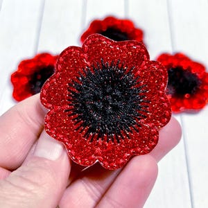 May include: Close-up of red glitter poppy flower embellishments with black centers. The petals have a scalloped edge. Several flowers are visible against a white background. These decorative flowers are suitable for crafts and embellishments.
