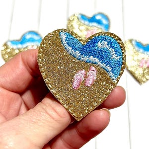 May include: Heart-shaped patch with a beach scene. The patch features a gold glitter background, blue embroidered waves, pink flip-flops, and a gold stitched border. Several similar patches are in the background.