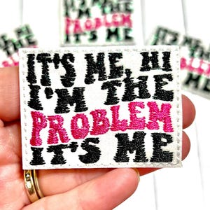 May include: White embroidered patch with the text "IT'S ME, HI, I'M THE PROBLEM IT'S ME" in black and pink. The patch has a stitched border and is likely an iron-on or sew-on patch for clothing or accessories.