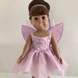 May include: A doll dressed as a fairy, wearing a light purple dress with matching wings. The doll has brown hair and is wearing a brown hair accessory. The dress has a bow at the waist and the wings are attached to the back.