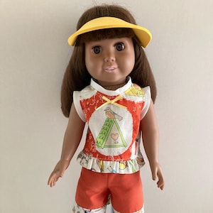 May include: A doll wearing a yellow visor, a white and orange top with a bird design, and orange shorts. The shorts and top have a patterned trim. The doll is also wearing sandals.