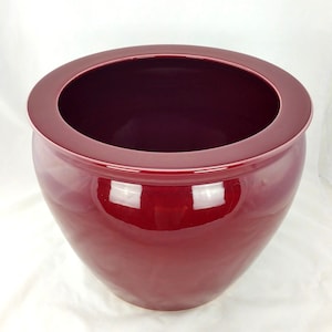 May include: A large, glossy, burgundy-colored ceramic planter with a wide, rounded rim. The planter has a bulbous shape, tapering towards the base. The interior is a darker shade of burgundy, creating a contrast with the exterior.
