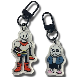 Può includere: Due portachiavi in acrilico trasparente con scheletri dei cartoni animati. Un portachiavi raffigura uno scheletro con una sciarpa e stivali rossi, l'altro mostra uno scheletro con una giacca blu e scarpe rosa. Entrambi i portachiavi hanno chiusure in metallo nero.