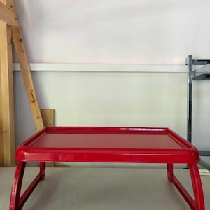May include: A bright red rectangular breakfast tray with a smooth, flat surface and a raised border edge. The tray features four foldable legs that extend outward, allowing it to stand independently on a flat surface for stable support.