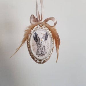 May include: A white egg-shaped ornament with a gold frame and a black and white image of a rabbit's face. The ornament is decorated with brown feathers and a beige ribbon.