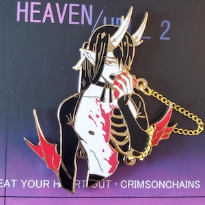 May include: Enamel pin featuring a demon-like character with horns, holding a red apple, and wearing a black and white outfit. The pin has a gold chain and red accents. Text on the card reads "HEAVEN / L 2" and "EAT YOUR HEART OUT - CRIMSONCHAINS."