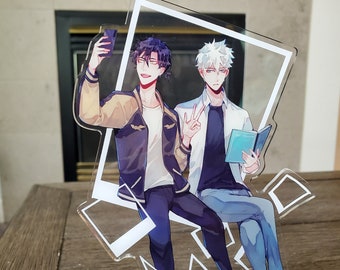 May include: A clear acrylic stand featuring two anime-style male figures. One figure is taking a selfie, while the other holds a book and makes a peace sign. The base has a clock design with Roman numerals.