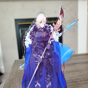 May include: A clear acrylic standee featuring a character with blonde hair, a blue cape, and armor. The character is holding a spear and standing on a blue base with red splatters. The standee is on a wooden surface.