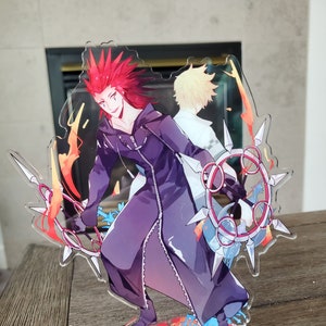 May include: A clear acrylic stand featuring two anime characters. The character on the left has red hair and is wearing a black and purple outfit. The character on the right has blonde hair and is wearing a white outfit. The stand has a red and gold base with a clock face and the word "Kingdom Hearts" written on it.
