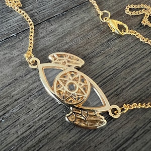 May include: A gold chain necklace with a pendant featuring a stylized eye with wings. The eye has a starburst design inside.