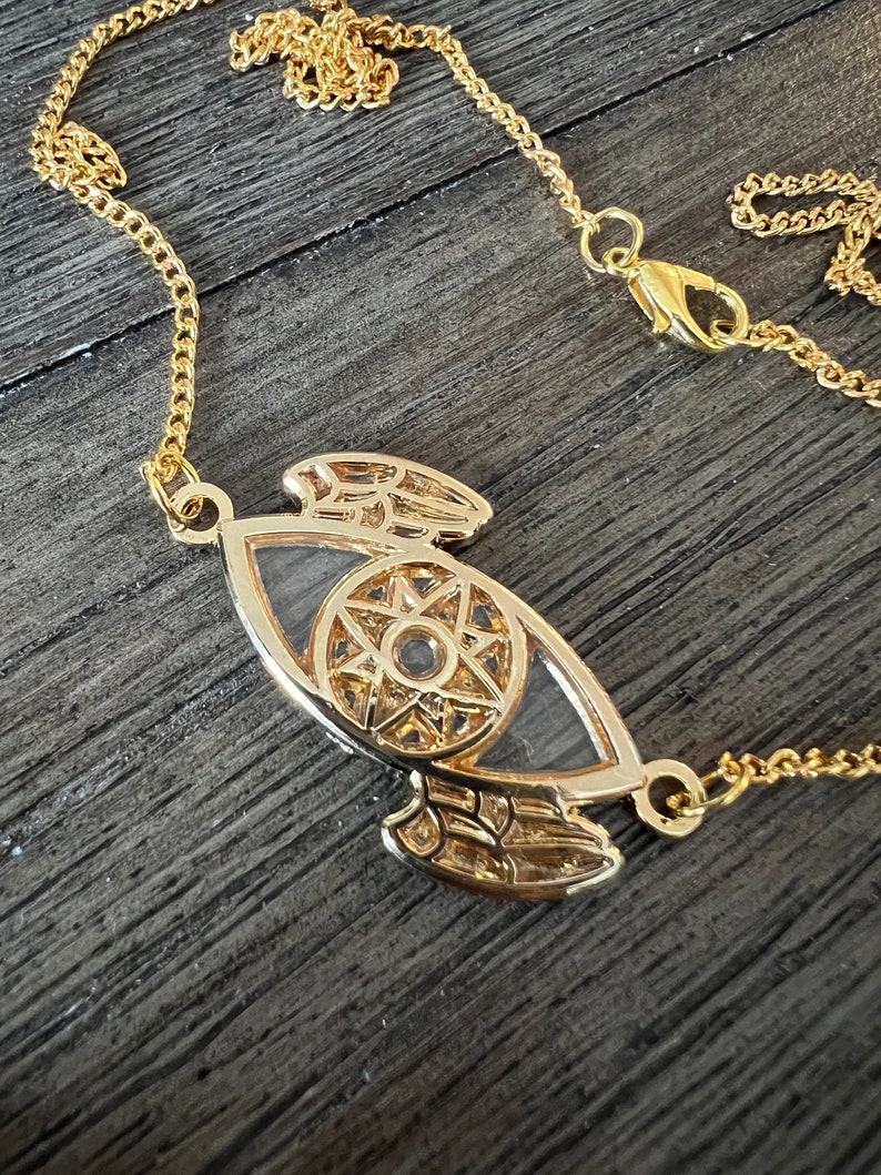 May include: A gold chain necklace with a pendant featuring a stylized eye with wings. The eye has a starburst design inside.