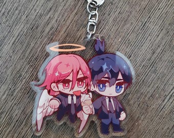 May include: A clear acrylic keychain featuring two chibi-style anime characters in dark suits. One character has pink hair, a golden halo, and white wings, holding an ice cream cone. The other character has dark blue hair and blue eyes.