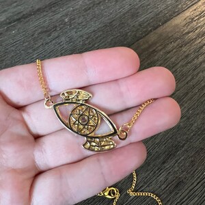 May include: A gold chain necklace with a pendant featuring a detailed eye design with a star inside. The eye is outlined in black and has a gold chain.