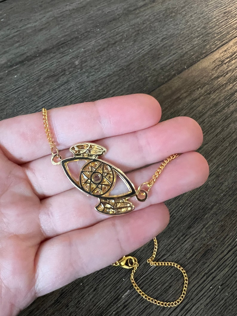 May include: A gold chain necklace with a pendant featuring a detailed eye design with a star inside. The eye is outlined in black and has a gold chain.