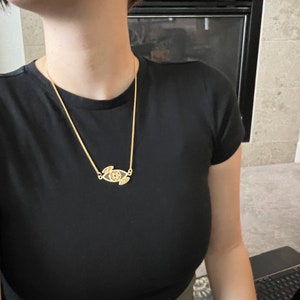 May include: A gold chain necklace with a pendant featuring a stylized eye design. The pendant is detailed with intricate lines and a small circle in the center.