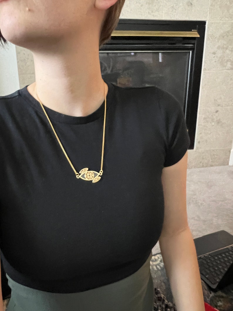 May include: A gold chain necklace with a pendant featuring a stylized eye design. The pendant is detailed with intricate lines and a small circle in the center.
