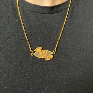 May include: A gold chain necklace with a black and gold eye pendant. The eye is detailed with intricate patterns.