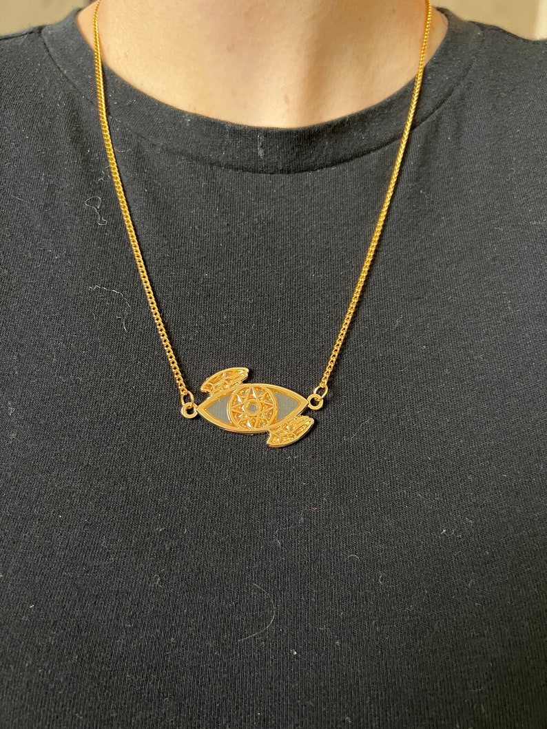 May include: A gold chain necklace with a black and gold eye pendant. The eye is detailed with intricate patterns.