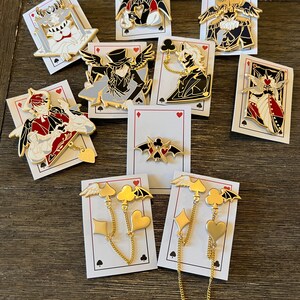 May include: A collection of enamel pins featuring various character designs, including winged figures and playing card suits. The pins are in gold, black, red, and white, displayed on playing card backings. Some pins have dangling chain accents.
