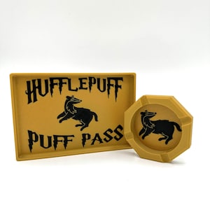 Może przedstawiać: Żółta tacka silikonowa z napisem "Hufflepuff Puff Pass" i czarnym wizerunkiem borsuka. Pasujący żółty popielniczkę silikonowa z czarnym wizerunkiem borsuka.