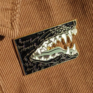 Può includere: Spilla rettangolare in smalto con un design dettagliato della bocca di un alligatore con denti bianchi affilati su sfondo nero. La spilla è attaccata a una camicia di velluto a coste marrone, con un bottone visibile. La spilla ha dettagli dorati e verdi.