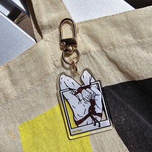 Puede incluir: Un llavero acrílico transparente con un cierre dorado. El llavero presenta un gráfico de un par de manos atadas con una cuerda roja, enmarcado en un borde blanco. El llavero cuelga de un bolso tote beige.