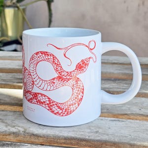 Op de afbeelding: Witte keramische mok met een rood slangenontwerp. De slangenillustratie wikkelt zich om de mok, met zijn tong uitgestoken. De mok heeft een gebogen handvat en de tekst "AVATAR DRAWS" is op de zijkant gedrukt.