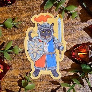 May include: A sticker of a black cat in knight's armour, holding a sword and shield. The shield has a sunburst design. The cat's armour is blue and red. The sticker is surrounded by green leaves and dice, on a wooden surface.