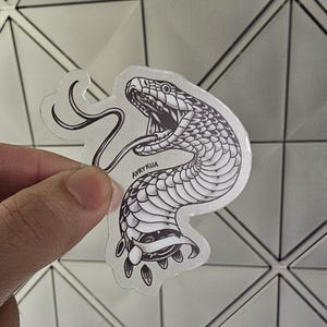May include: A black and white sticker of a snake with its mouth open and tongue out. The snake is coiled and has a detailed pattern of scales. The sticker is on a clear background and has the text "AVRYKUA" printed on it.