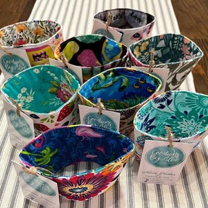 May include: A collection of colourful fabric coffee cup sleeves with a variety of patterns, including floral, geometric, and abstract designs. Each sleeve has a white tag with the text "Creations By Alice" and a handwritten note.