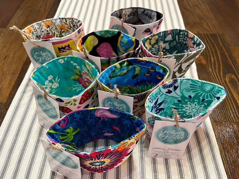 May include: A collection of colourful fabric coffee cup sleeves with a variety of patterns, including floral, geometric, and abstract designs. Each sleeve has a white tag with the text "Creations By Alice" and a handwritten note.