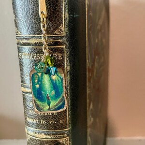 May include: A gold-toned bookmark with a chain and a rectangular charm featuring a green and blue illustration of a person in a forest. The bookmark is attached to a vintage book with the title "SHAKESPEARE".