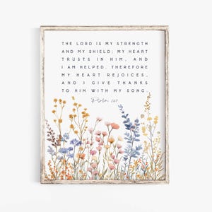 May include: A framed wall art print featuring a white background with black text that reads: The Lord is my strength and my shield; my heart trusts in him, and I am helped. Therefore my heart rejoices, and I give thanks to him with my song. Psalm 28:7. The bottom displays watercolor wildflowers in shades of yellow, blue, pink, and orange.