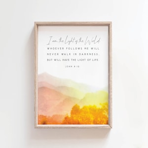 May include: A framed print with a watercolour background of a mountain range in shades of yellow, orange, and pink. The print features a quote from the Bible, John 8:12, "I am the Light of the World. Whoever follows me will never walk in darkness, but will have the light of life."