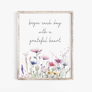 Puede incluir: Una impresión de acuarela con fondo blanco y una cita que dice "begin each day with a grateful heart". La cita está escrita en letra cursiva negra. La impresión presenta un borde de flores de acuarela en tonos de rosa, morado y amarillo.