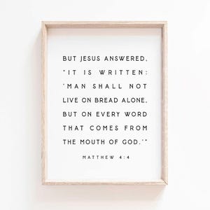 Op de afbeelding: Een ingelijste print met een licht houten frame. De print bevat zwarte tekst op een witte achtergrond, met een citaat uit Mattheüs 4:4: "Maar Jezus antwoordde..." Het citaat gaat verder met de woorden "De mens zal niet van brood alleen leven..."