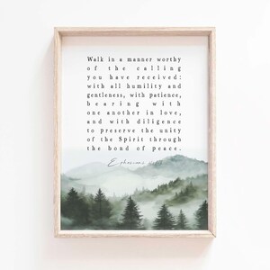 May include: A framed print featuring a serene landscape of misty green mountains and evergreen trees. The artwork includes a quote from Ephesians 4:1-3 in black text. The frame is a light wood color, adding a natural touch to the decor.