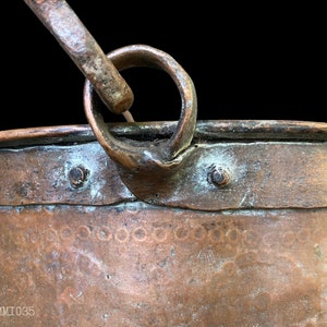 May include: Close-up of a copper bucket with a metal handle. The bucket has a hammered finish and is secured with rivets. The handle is attached to the bucket with a metal ring.