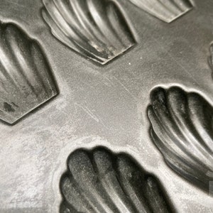 May include: A black metal baking pan with multiple individual moulds shaped like seashells. The moulds are designed for making madeleines.