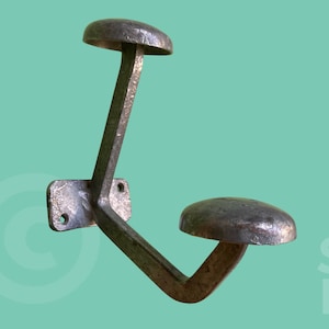 Peut inclure: Un crochet mural en métal de style vintage avec deux protubérances arrondies en forme de chapeau. Le crochet est de couleur gris foncé vieilli et est monté sur une plaque rectangulaire avec deux trous de vis. Les mots "BRIQUE" et "STUDIO" sont visibles.