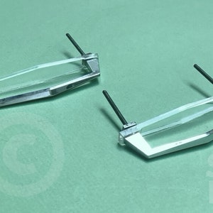 Op de afbeelding: Twee zilverkleurige kastdeurgrepen met heldere glazen inzetstukken en zwarte schroeven. De grepen hebben een geometrische vorm.