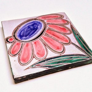 May include: A square ceramic tile with a hand-painted design of a pink flower with a blue center. The flower has a gold outline and is surrounded by green leaves.