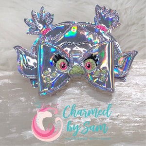 May include: A holographic silver bow with a green and pink embroidered face and paw prints. The bow has two pigtails on top.