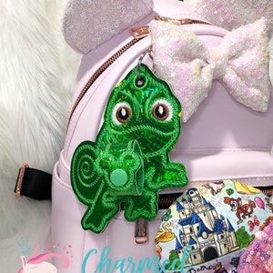 May include: A pink faux leather backpack with a green chameleon keychain and a pink and white sequin bow. The backpack has a zipper closure and a gold zipper pull. The keychain is attached to a gold key ring. The bow is attached to the top of the backpack. The backpack is sitting on a white surface.