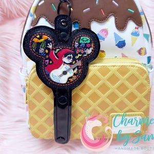Puede incluir: Llavero negro con forma de Mickey Mouse con un diseño bordado colorido de la película Coco. El llavero está sujeto a una correa negra y se coloca contra una mochila con temática de postres y una cartera amarilla con estampado de gofres.