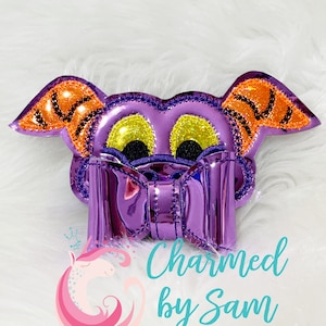 May include: Purple and orange felt character with large eyes and a purple bow. The character is a felt applique for a hair bow or other craft project. "Charmed by Sam" is written below the character.