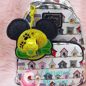 May include: A white backpack with a black and green Mickey Mouse charm. The backpack has a pattern of dog houses and dogs. The charm has glitter and a green bow. The backpack has the text "Disney" on it.